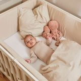 Baby in einem cremefarbenen FLEXA-Babybett (140 × 70 cm) mit beiger Decke und Plüsch-Krake, abgebildet in einem Kinderzimmer.