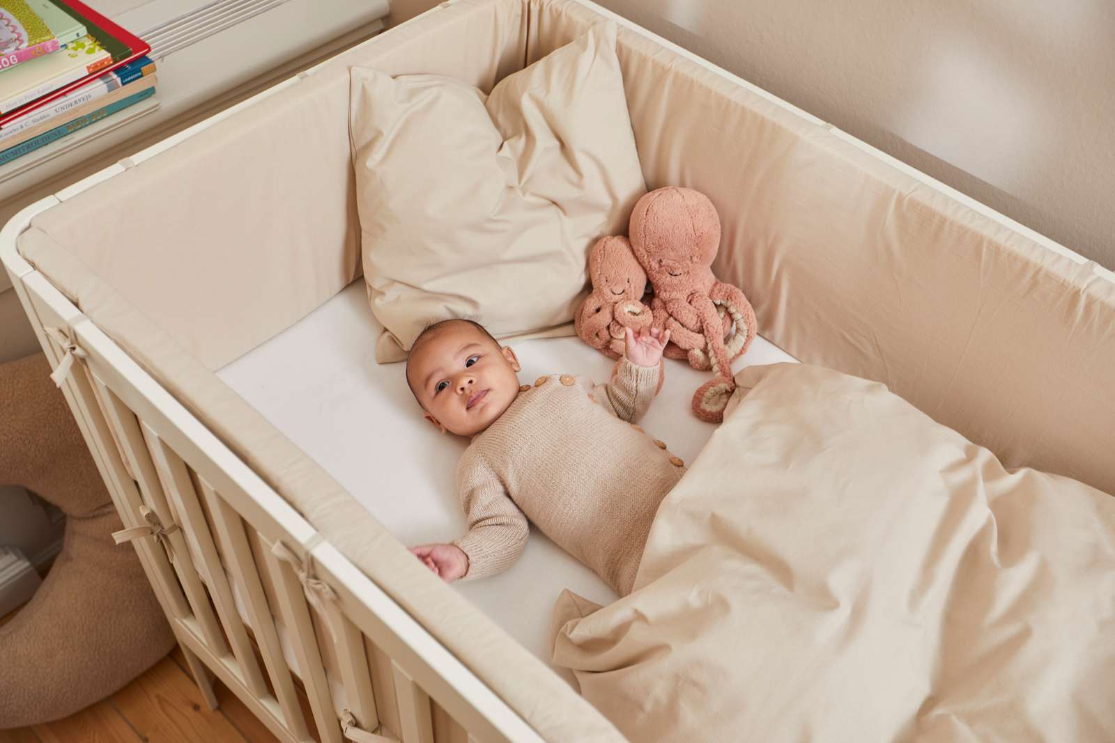 Baby in einem cremefarbenen FLEXA-Babybett (140 × 70 cm) mit beiger Decke und Plüsch-Krake, abgebildet in einem Kinderzimmer.