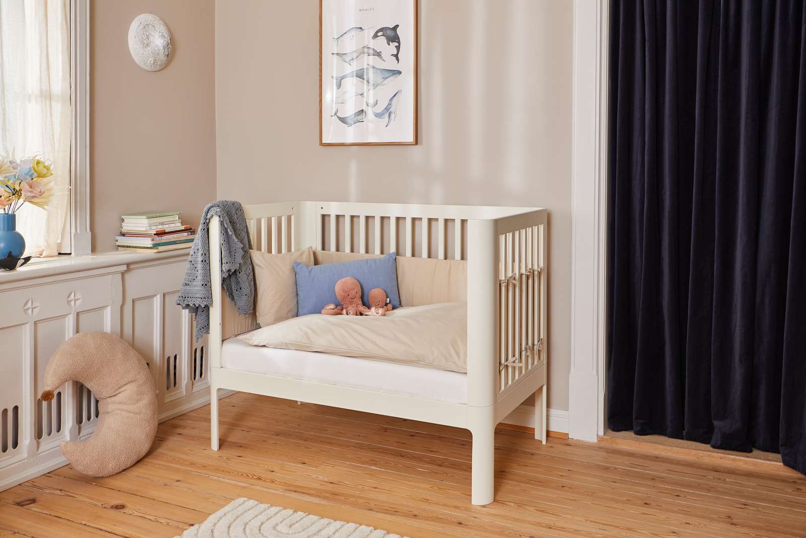 Cremefarbenes FLEXA-Babybett, 140 x 70 cm, aus Holz, mit Kissen und Spielzeug in einem sanft beleuchteten Kinderzimmer mit kindersicheren, abgerundeten Kanten.