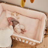 Femme à côté dun lit bébé FLEXA de la collection OAK, finition bois clair, dans une chambre denfant où lon aperçoit les bords arrondis, sans danger pour les enfants.