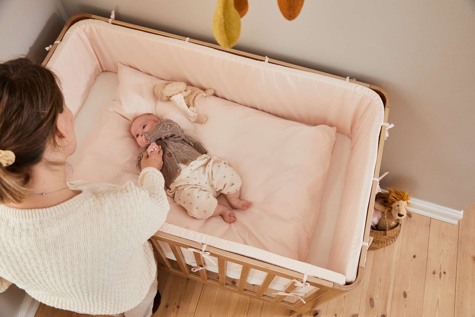 Frau neben einem FLEXA-Babybett aus der OAK-Kollektion, helle Holzoptik, im Kinderzimmer mit kindgerechten, abgerundeten Kanten.