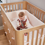Bébé avec une tétine dans un lit bébé FLEXA, en chêne, avec literie moelleuse et tours de lit, chambre denfant ; barrières de sécurité visibles.