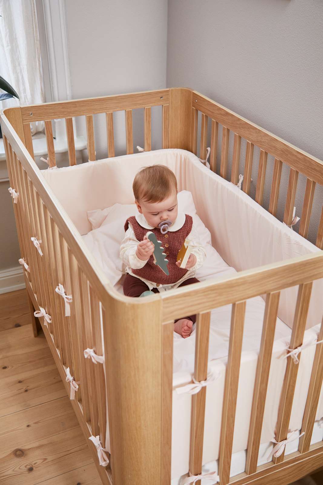 Bébé avec une tétine dans un lit bébé FLEXA, en chêne, avec literie moelleuse et tours de lit, chambre denfant ; barrières de sécurité visibles.