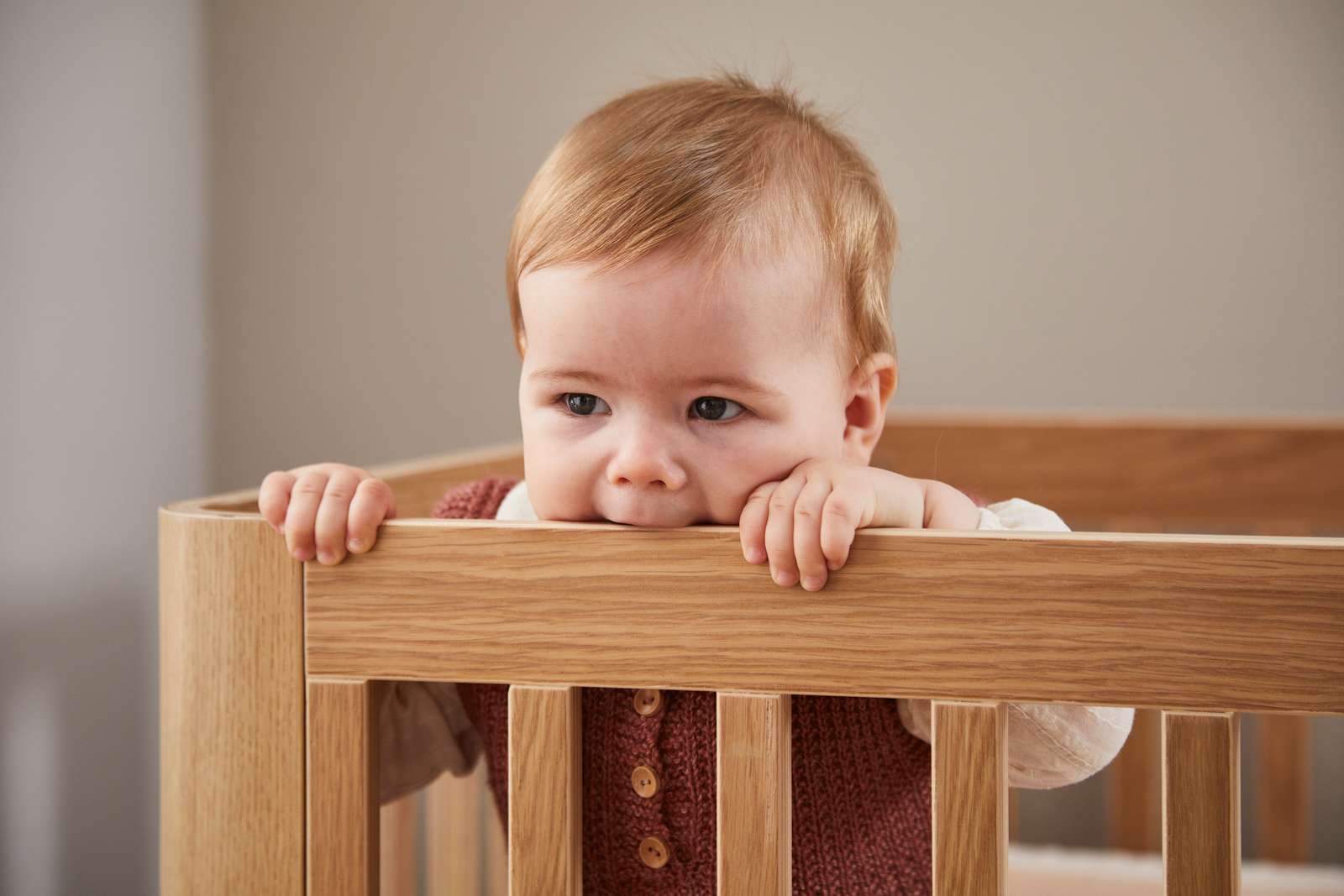 Bébé dans un lit bébé FLEXA OAK, mordillant sans danger la barre supérieure ; conception sécurisée pour les enfants, finition chêne, décor de chambre denfant visible.