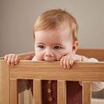 Bébé dans un lit bébé FLEXA OAK, mordillant sans danger la barre supérieure ; conception sécurisée pour les enfants, finition chêne, décor de chambre denfant visible.