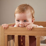 Bébé dans un lit bébé FLEXA OAK, mordillant sans danger la barre supérieure ; conception sécurisée pour les enfants, finition chêne, décor de chambre denfant visible.